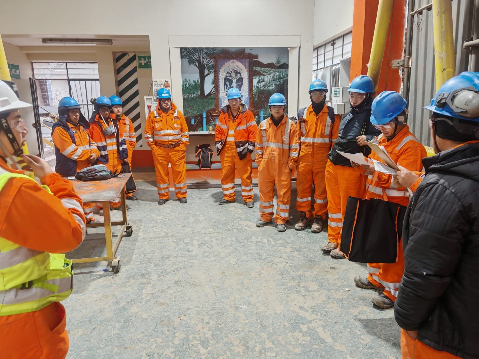 Charla de seguridad con un grupo de trabajadores con overol naranja y casco azul dentro de una planta