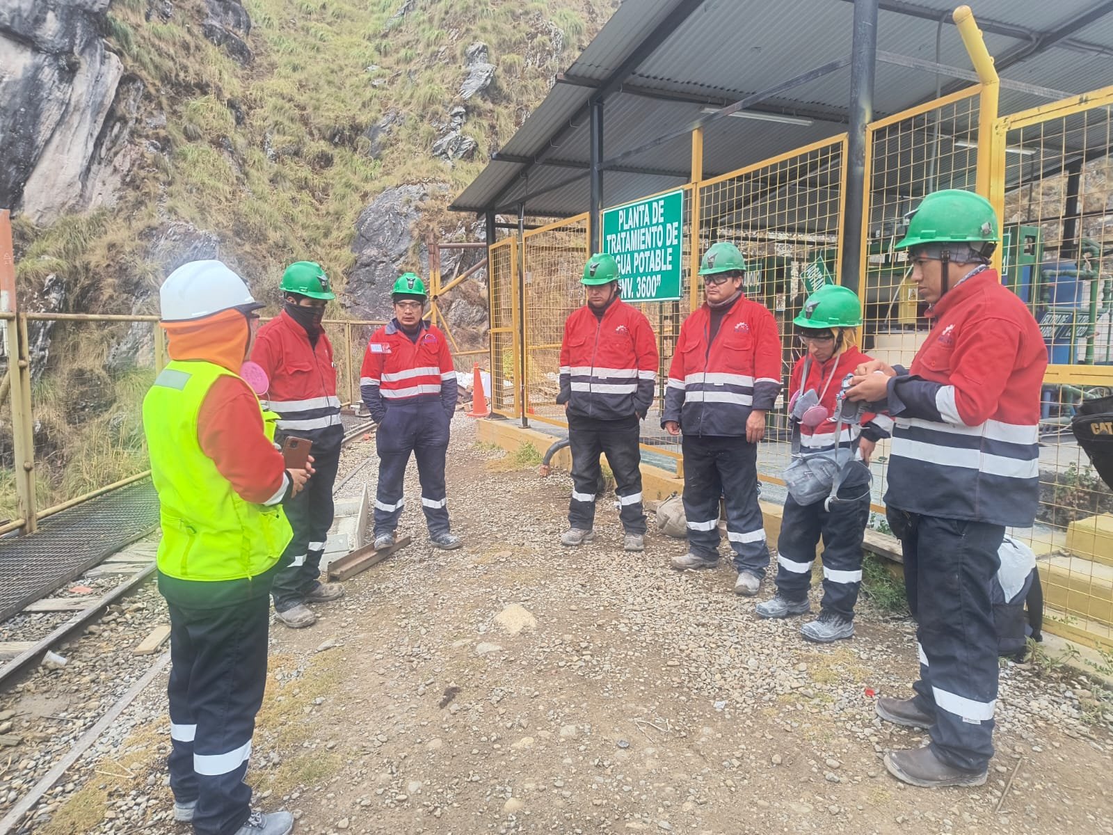 Charla de seguridad con un grupo de trabajadores con casco verde frente a una planta de tratamiento de agua en zona minera